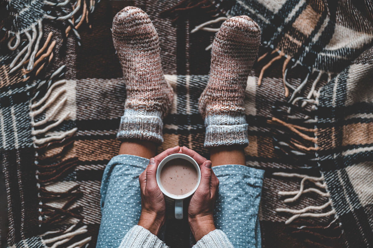 A top-down shot of a pair of hands holding hot cocoa in their lap while wearing fuzzy socks and sitting on a brown plaid blanket.